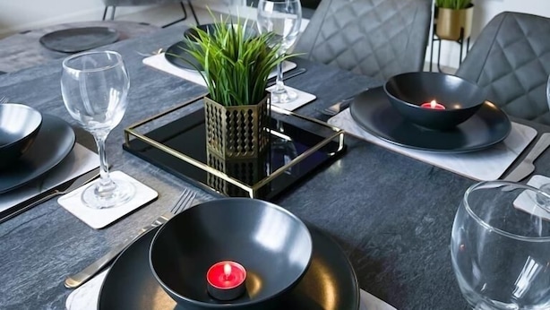 Un comedor moderno con una mesa negra dispuesta para cuatro personas, un televisor montado en la pared y un sofá gris al fondo.