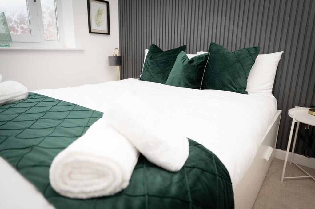 A neatly made bed with a green quilt and white pillows, a small bedside table, and a window with a view of trees.