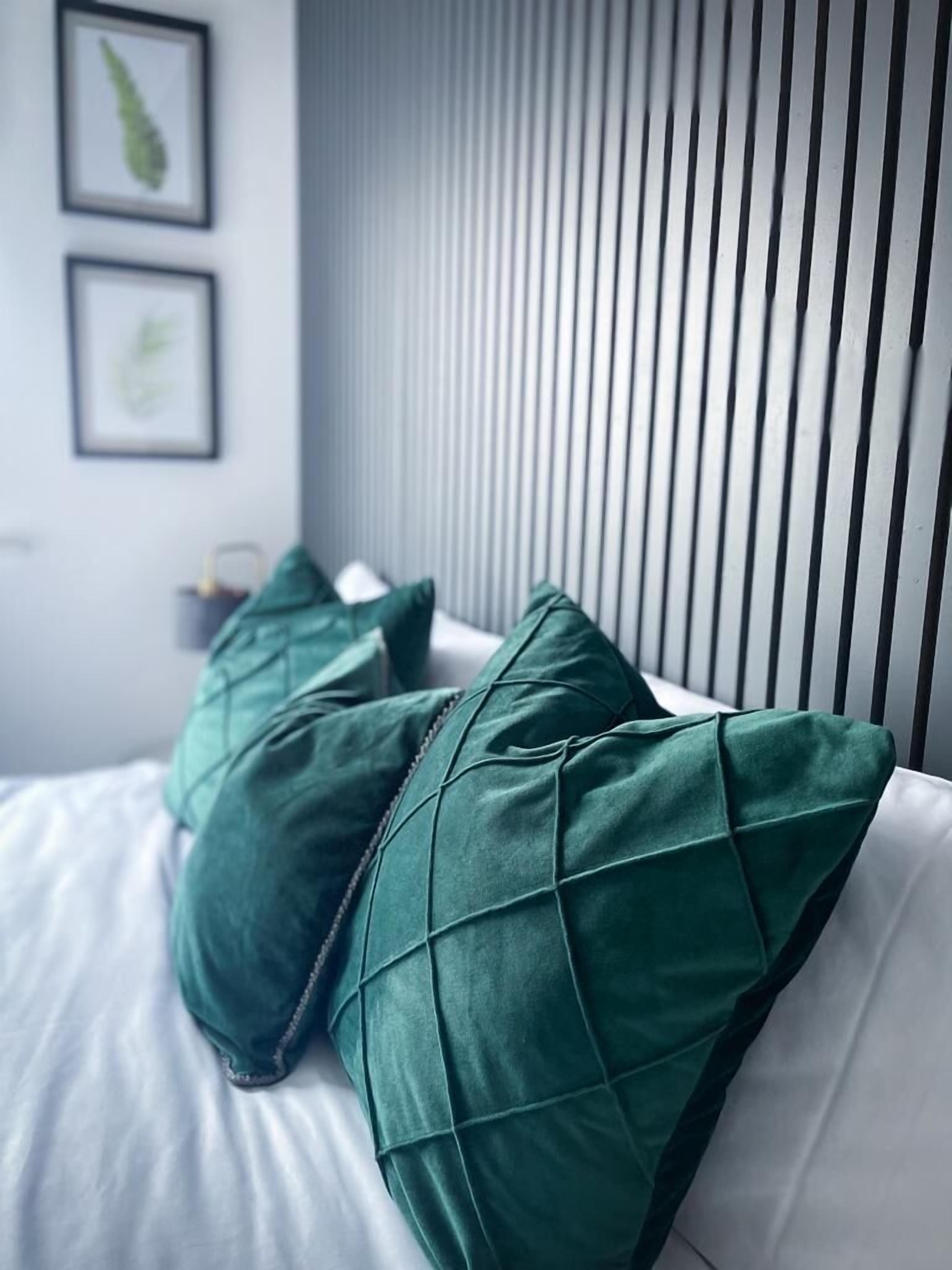 Llits amb almofades verdes i un panell de capçal blanc en una habitació moderna amb persianes verticals.