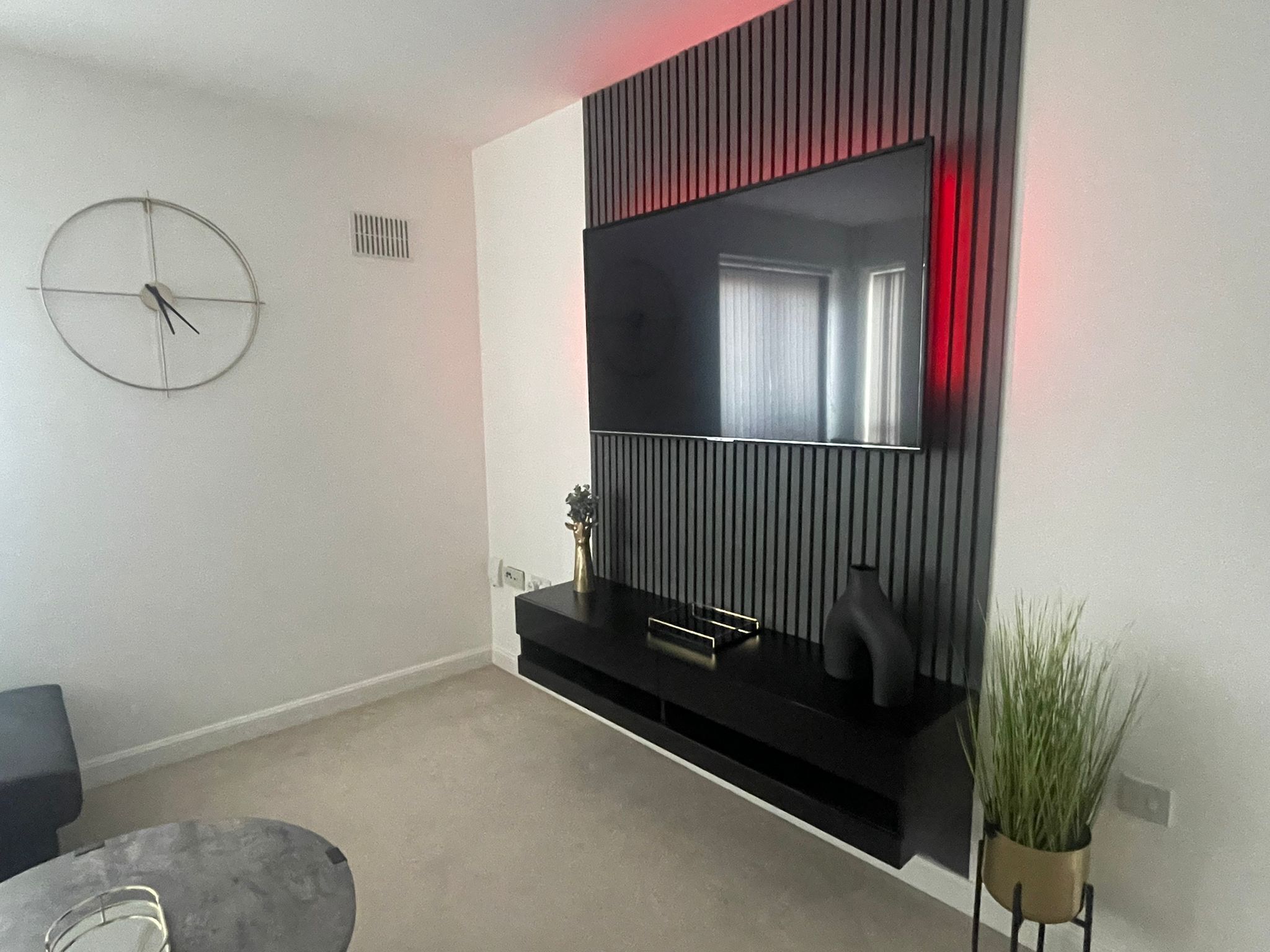 A modern living room with a large flat-screen TV mounted on a black wall, a round clock, a small plant, and a grey sofa.