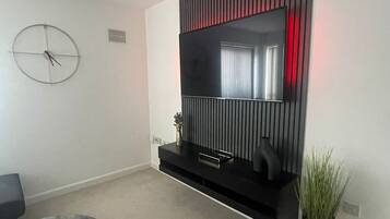 A modern living room with a large flat-screen TV mounted on a black wall, a round clock, a small plant, and a grey sofa.