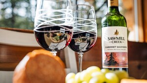 Two wine glasses with a bottle of Sawmill Creek Cabernet Sauvignon in the foreground, with a blurred background of greenery. - Pet-Friendly Off-grid Log Cabin, Hiking, Mountain Air and Dark Skies Await! (Rock Lake Provincial Park)