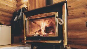 A wood-burning stove in a log cabin with a visible fire, a white appliance in the background, and a woven mat on the floor. - Pet-Friendly Off-grid Log Cabin, Hiking, Mountain Air and Dark Skies Await! (Rock Lake Provincial Park)