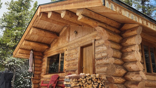 A log cabin with a wooden porch, a stack of firewood, and a red chair. - Pet-Friendly Off-grid Log Cabin, Hiking, Mountain Air and Dark Skies Await! (Rock Lake Provincial Park)