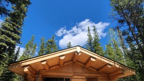 A log cabin with a covered patio, two Adirondack chairs, and a table with a grill. - Pet-Friendly Off-grid Log Cabin, Hiking, Mountain Air and Dark Skies Await! (Rock Lake Provincial Park)