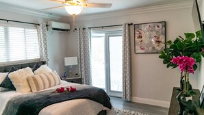 A bedroom with a bed, a ceiling fan, a nightstand, a lamp, a window with blinds, a door, and a wall-mounted air conditioner. - Entire Condominium Home away from Home with Pool, Near Beach (Nassau)