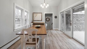 A dining area with a wooden table and white chairs, a chandelier, and large windows. - Updated Biddeford Pool Beach House (Biddeford)