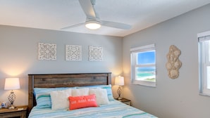 Une chambre à coucher avec un lit, un ventilateur de plafond, des œuvres d’art décoratives au mur et une fenêtre donnant sur un paysage agréable.