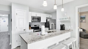 A modern kitchen with white cabinetry, a granite countertop, a built-in microwave, and a stainless steel refrigerator. There are white bar stools and pendant lights above the counter. - 3BR @ Grand Beach Resort • Sleeps 8 • Balcony, Pool • Walk to Beach & The Hangout (Gulf Shores)