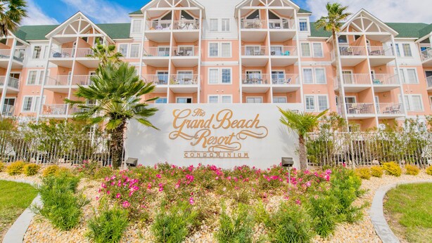 A multi-story building with balconies, a sign reading "The Grand Beach Resort Condominium," and landscaped gardens with various plants and flowers. - 3BR @ Grand Beach Resort • Sleeps 8 • Balcony, Pool • Walk to Beach & The Hangout (Gulf Shores)