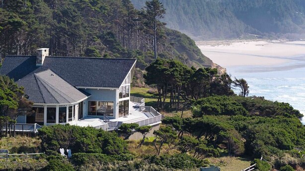 Een huis aan de kust met een groot terras, met uitzicht op een strand en beboste heuvels.