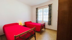Una habitación con dos camas individuales, una silla de madera, una mesita con una lámpara y una ventana con cortinas estampadas.