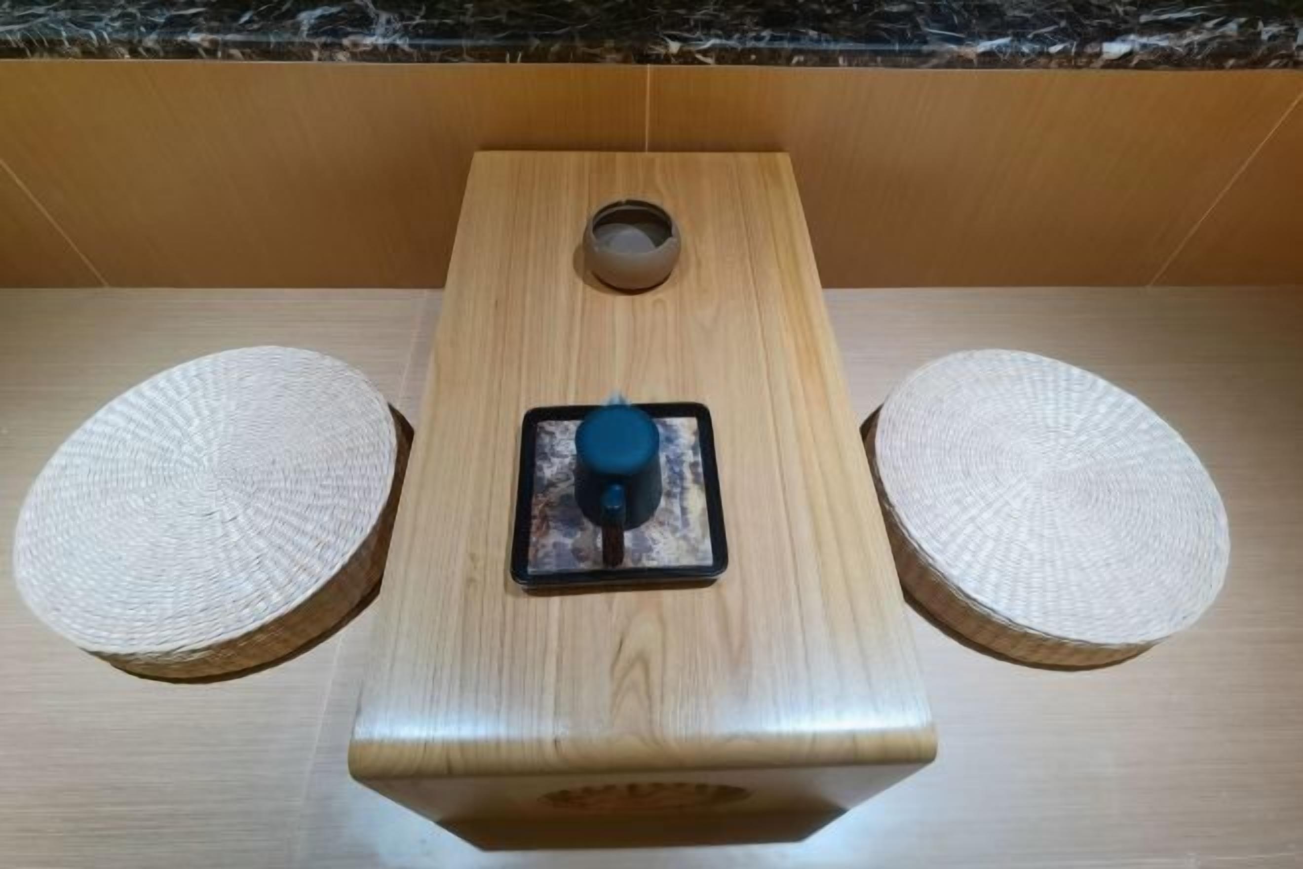 A wooden table with a blue object in the center, two woven stools, and a wooden panel with a circular cutout.