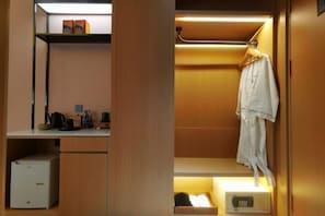 A compact hotel room with a kitchenette, a robe hanging on the wall, and a small bedside table with a lamp.