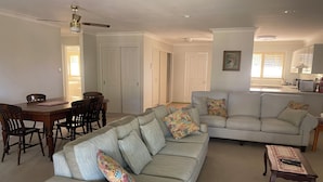A living room with a sofa, a dining table with chairs, a ceiling fan, and a kitchen area in the background. - 2 bedroom 2 bathroom apartment (Mollymook)