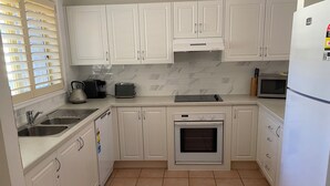 A kitchen with white cabinets, a built-in oven, a microwave, a sink, and a window with blinds. - 2 bedroom 2 bathroom apartment (Mollymook)