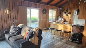 A cozy cabin interior with a wooden ceiling, a sofa with patterned cushions, a kitchen area with stools, and a large window providing a view of the outdoors. - Upper Red Lake all season destination!  (Waskish)