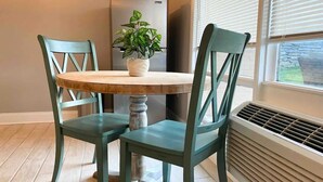 A dining area with a round table and two chairs, a potted plant, a refrigerator, and a window with blinds.