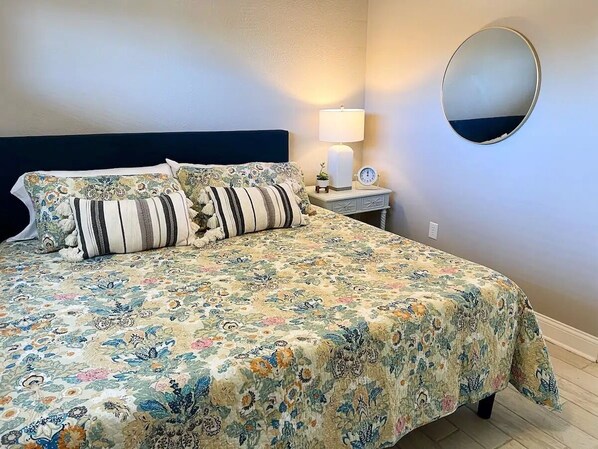 Un lit bien fait, avec une literie à motifs floraux, une lampe de chevet et un miroir rond accroché au mur.