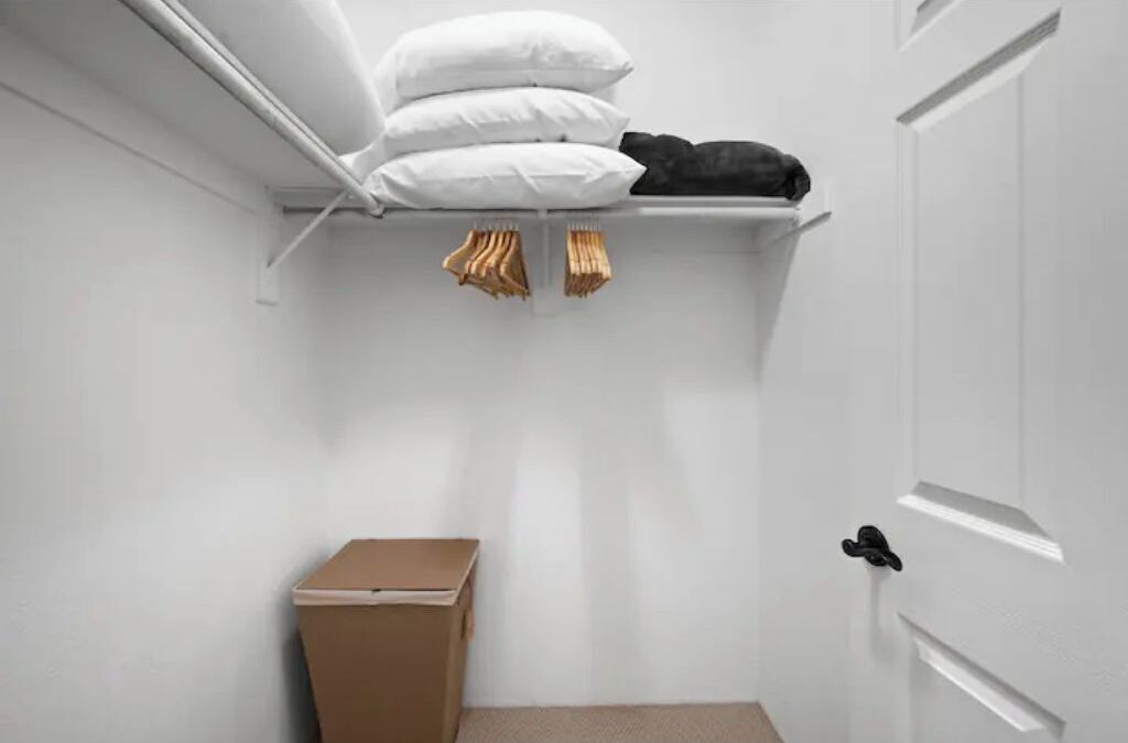 Una habitación pequeña con paredes blancas, una puerta, un estante blanco con almohadas y una chaqueta negra, además de una caja de cartón en el suelo.