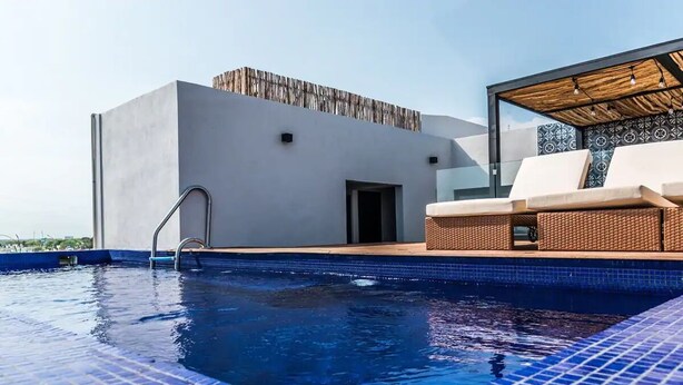 Une piscine sur le toit offrant une vue dégagée sur le ciel, un bâtiment moderne et un salon aménagé avec des meubles en osier.
