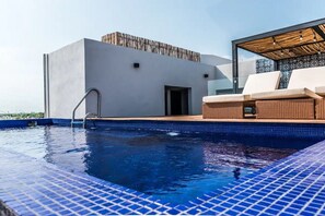 Une piscine sur le toit offrant une vue dégagée sur le ciel, un bâtiment moderne et un salon aménagé avec des meubles en osier.