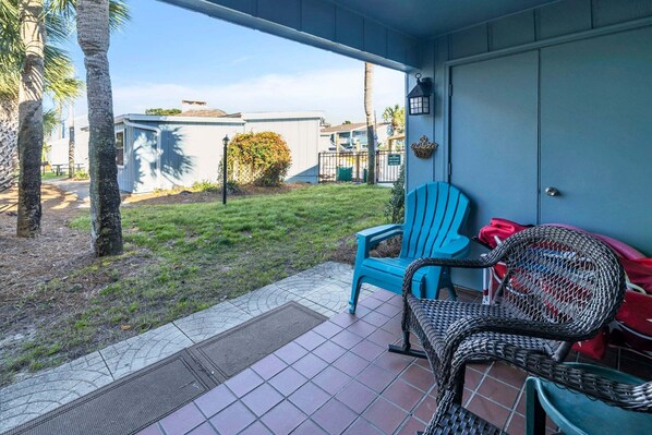 A covered patio area with a tiled floor, two chairs, a wicker chair, and a small garden with grass and a tree. - Beach Service Included, Pet Friendly! Pickle Ball Courts , CLM 11A! (Destin)