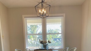 Un coin repas avec une table en bois, des chaises blanches, un lustre et une vue par la fenêtre.