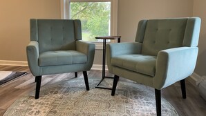 Deux fauteuils bleu clair aux pieds noirs, un tapis à motifs et une petite table dans une pièce dotée d’une fenêtre et de murs de couleur beige.