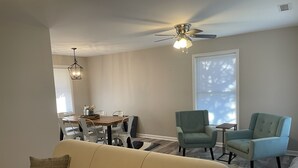 Un salon avec un canapé beige, deux fauteuils, une table à manger avec des chaises, un ventilateur de plafond et une fenêtre avec des stores.