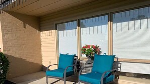A covered patio with two blue chairs and a table, a potted plant with flowers, and a brick wall. - Capital City Condo #4 (Frankfort)