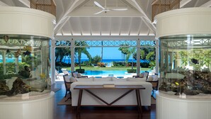 A spacious room with a large glass fish tank, a pool visible through the windows, and a ceiling fan. - The Great House | Beach Front - Located in  Exquisite St. Peter with Private Pool (Mullins)
