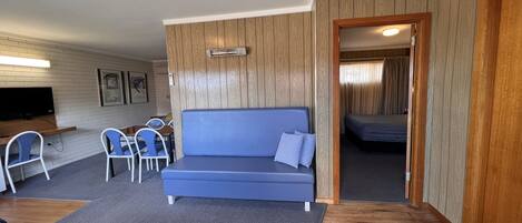 A compact living space with a blue sofa, a small dining area with chairs, a television, and a wooden accent wall.