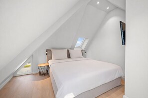Un dormitorio con techo inclinado, una cama, una mesita de noche, una lámpara y una ventana.