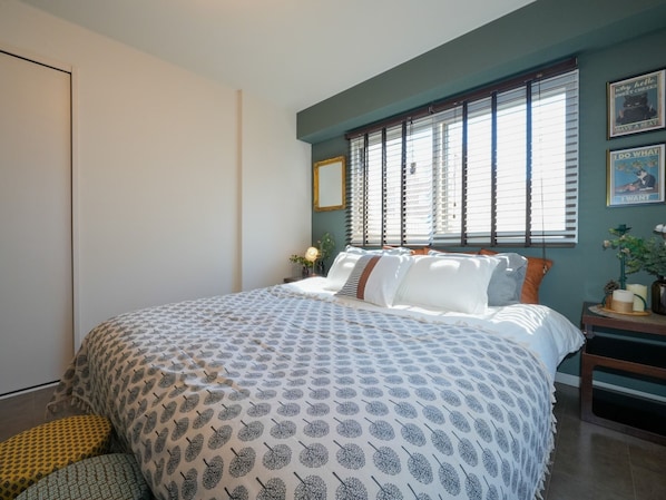 A bedroom with a large bed, a window with blinds, a bedside table with a lamp, and a wall with framed pictures.