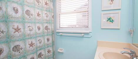A bathroom with a seashore-themed shower curtain, a window with blinds, and framed artwork on the wall.
