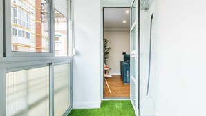 Un couloir étroit doté d’un sol recouvert d’un gazon artificiel vert, une porte donnant sur une pièce où se trouvent un canapé bleu et une petite table, ainsi qu’une fenêtre donnant sur un immeuble.