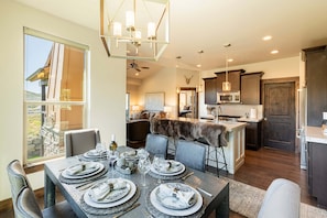 A dining area with a table set for six, a kitchen with dark wood cabinets, and a view of a mountainous landscape through a large window. - Luxury Townhome, Theater & Family Room, Hot Tub! 2 Masters (Kamas)