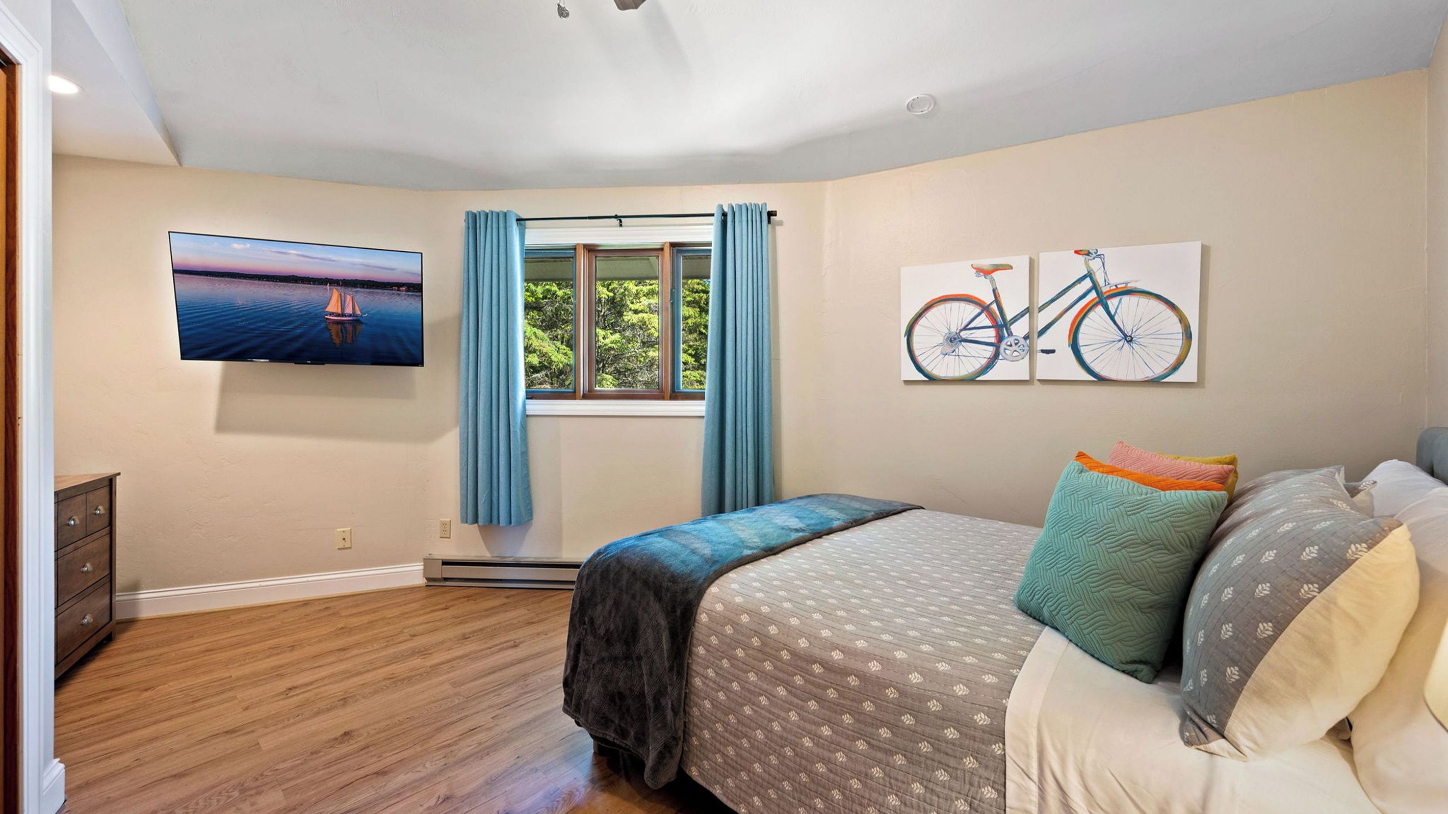 Un dormitorio con cama, televisión, ventana con cortinas y dos cuadros colgados en la pared.