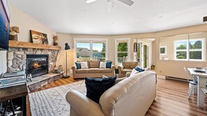 A cozy living room with a stone fireplace, comfortable seating, and large windows offering a view of the outdoors.