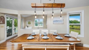 A dining room with a wooden table set for four, white chairs, and a view of the outdoors through large windows.