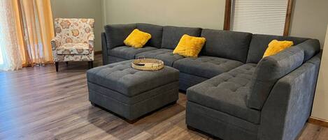 Una sala de estar con un sofá seccional gris, dos cojines amarillos y un reposapiés gris con una bandeja trenzada.
