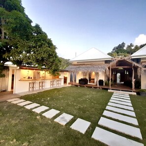 Une maison moderne dotée d’un espace extérieur aménagé avec un coin salon couvert, d’une cuisine et d’une pelouse bien entretenue.