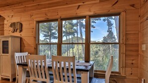 Interior de uma cabana de madeira com uma mesa de jantar e cadeiras, uma janela com vista para a floresta e uma decoração em forma de urso na parede.
