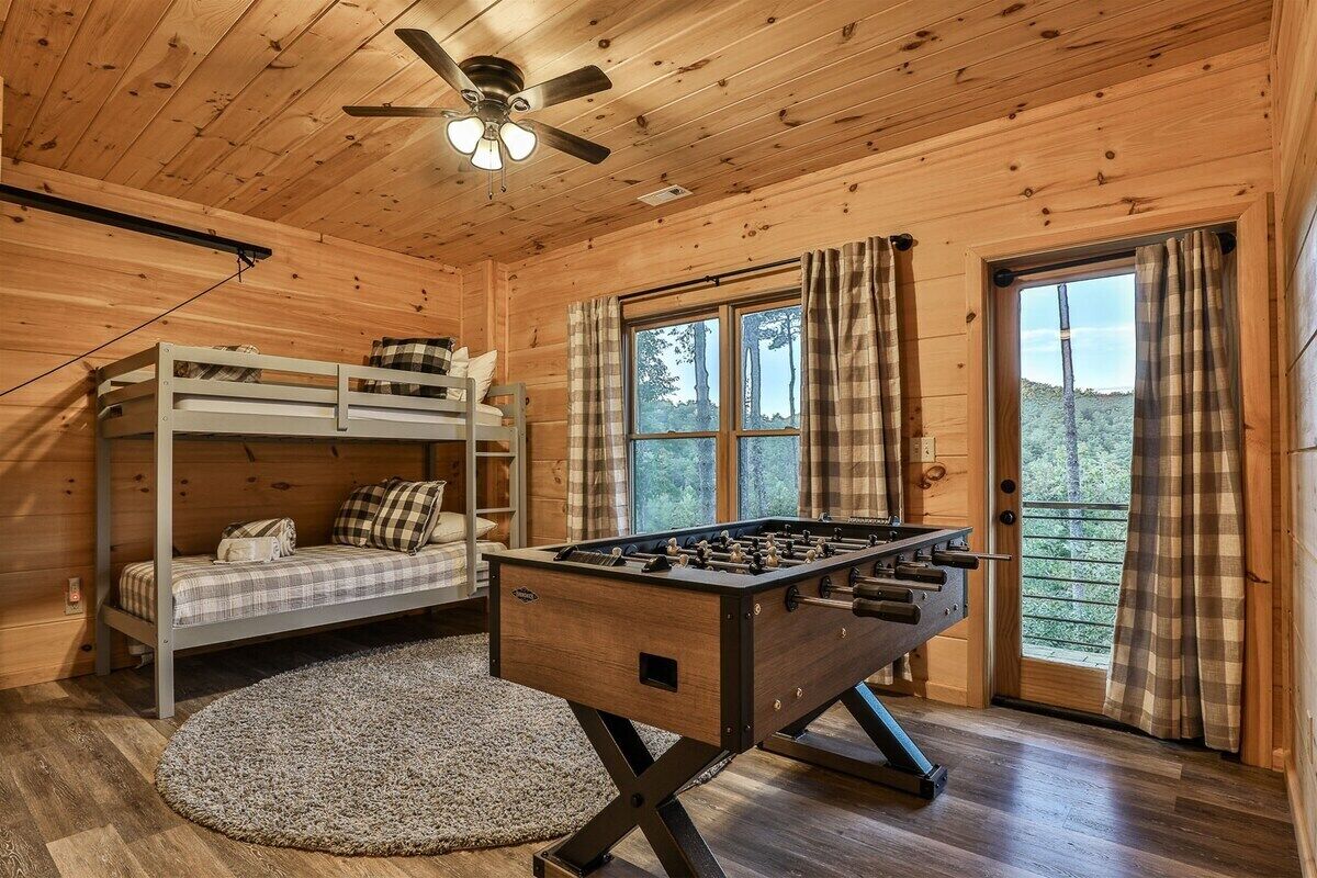 A wooden cabin interior with a bunk bed, a foosball table, a ceiling fan, and a window with curtains.