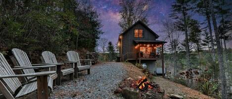 A cabin with a deck, a fire pit, and a starry night sky.
