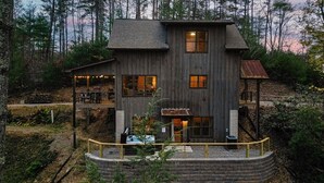 Uma cabana de madeira com dois andares, um deque coberto, uma área externa de estar e uma estrutura circular de pedra na base.