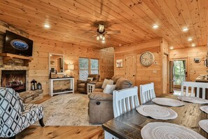 A log cabin interior with a fireplace, seating area, dining table, and a clock on the wall.
