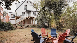 Un jardin avec des fauteuils Adirondack et un foyer, entouré d’une maison blanche dotée d’un balcon.