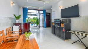 A modern hotel lobby with a wooden dining table, chairs, a potted plant, a large flat-screen TV, and a sound system. - Gió Chiều Homestay - Riverside & Swimming pool (Hội An)